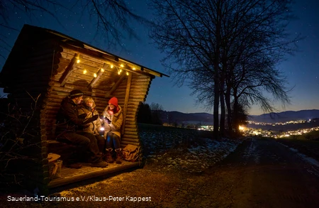 Drei Menschen sitzen im Unterstand im Dunkeln, in der Ferne leuchten die Lichter eines Dorfes