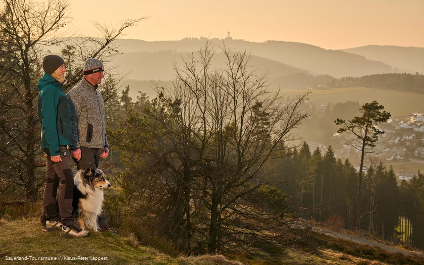 Ein Wanderpaar mit Hund im Abendlicht im Hintergrund der Ettelsberg