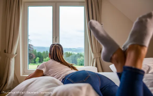 Frau schaut auf dem Bett leigend aus dem Fenster in die Landschaft