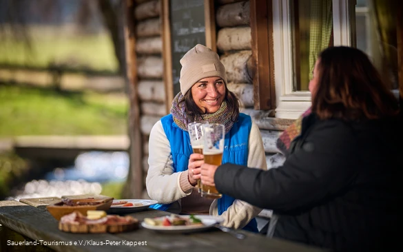 Eine Frau mit Mütze stößt mit einer andern Frau mit Bier an. Beide sitzen vor einer Hütte