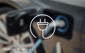 Ein verschwommenes Bild eines Ladesteckers in einer Autoladeklappe mit der Zeichnung eines Haushaltsteckers im Vordergrund. Dieses Bild dient als Symbolbild für E-Car Ladestationen.