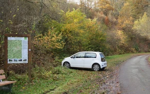 Ein Parkplatz auf dem ein kleines weißes Auto parkt neben einem Waldweg mit einer Infotafel zu Wanderwegen.