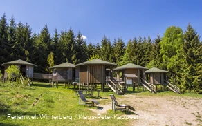 Fünf hölzerne, runde Hütten auf Stelzen mit jeweils einer Treppe davor umgeben von Wald und Wiesen.