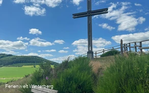 Ein großes Holzkreuz auf einem begrünten Hügel, davor eine Bank.