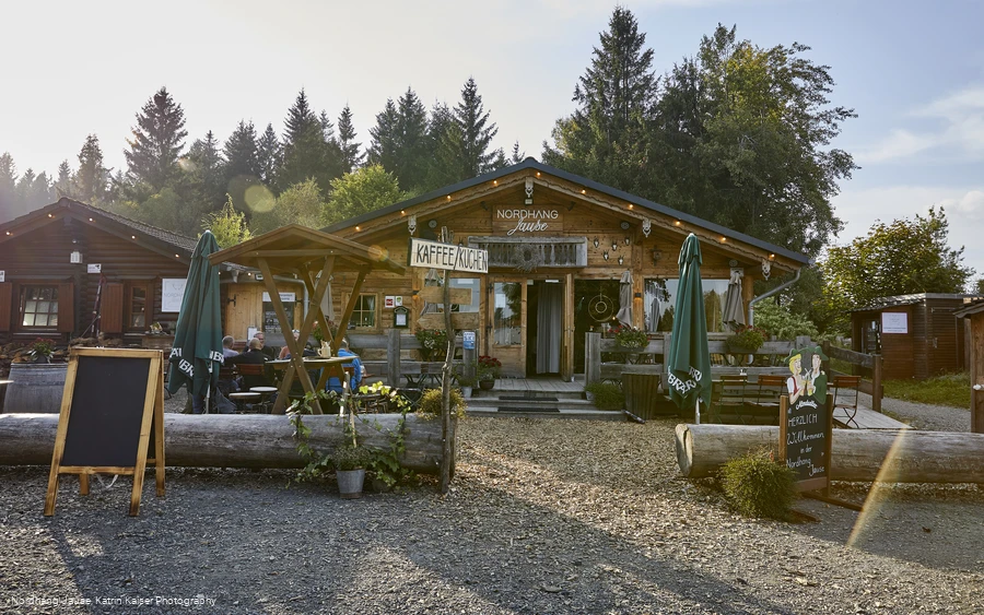 Außenansicht einer hölzernen Hütte mit der Aufschrift Nordhang Jause mit einigen Sitzmöbeln und Schildern vor der Tür.