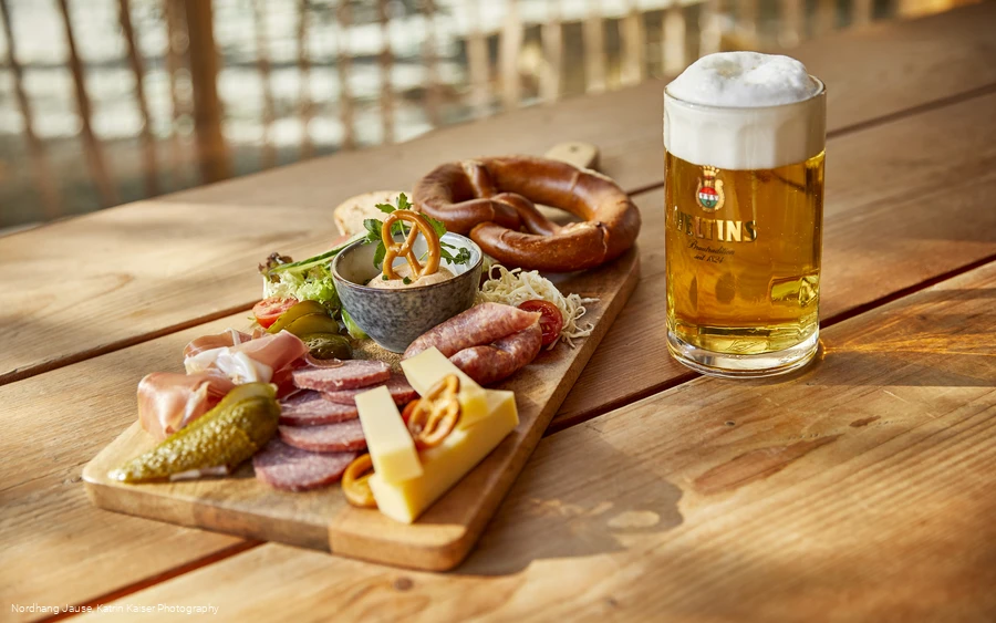 Ein hölzernes Brett mit einigen Speisen neben einem gefüllten Bierkrug auf einem Tisch.
