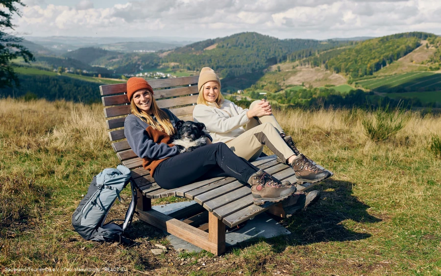 Zwei Frauen und ein Hund machen eine Pause auf einer Bank mit weiter Aussicht