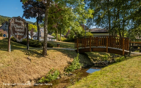 Eine hölzerne Brücke über einem Bach umgeben von Wiese und Bäumen und einem Holzschild mit der Aufschrift Besuchen Sie Elkeringhausen im Orketal.