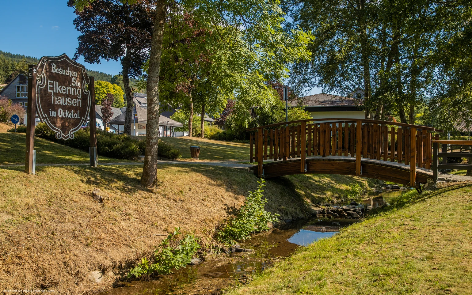 Eine hölzerne Brücke über einem Bach umgeben von Wiese und Bäumen und einem Holzschild mit der Aufschrift Besuchen Sie Elkeringhausen im Orketal.
