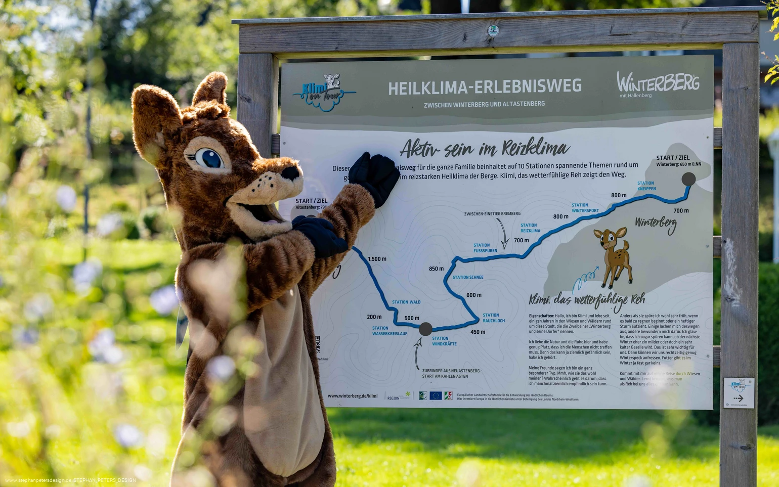 Waldi zeigt auf das Heiklima-Erlebnisweg-Schild
