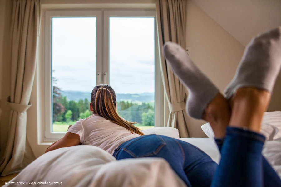 Frau schaut auf dem Bett leigend aus dem Fenster in die Landschaft