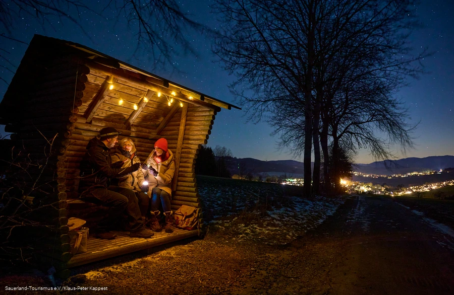Drei Menschen sitzen im Unterstand im Dunkeln, in der Ferne leuchten die Lichter eines Dorfes
