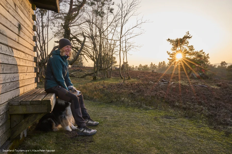 Auf einer Bank an einer Hütte auf dem Kahlen Pön sitzt Frau mit Hund im Winterlicht