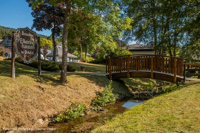Eine hölzerne Brücke über einem Bach umgeben von Wiese und Bäumen und einem Holzschild mit der Aufschrift Besuchen Sie Elkeringhausen im Orketal.