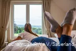 Frau schaut auf dem Bett leigend aus dem Fenster in die Landschaft