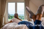 Frau schaut auf dem Bett leigend aus dem Fenster in die Landschaft