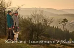 Ein Wanderpaar mit Hund im Abendlicht im Hintergrund der Ettelsberg