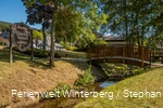 Eine hölzerne Brücke über einem Bach umgeben von Wiese und Bäumen und einem Holzschild mit der Aufschrift Besuchen Sie Elkeringhausen im Orketal.